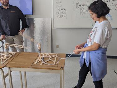 Female student focuses on built design for engineering class, male teacher watches