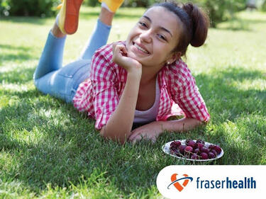 A young girl smiling while laying on the grass. The Fraser Health logo is located in the bottom right corner